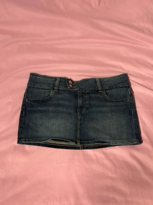 H&M jeanskjol  - Trendig jeanskjol från H&M i kort mini-modell med Y2K-känsla. Klassisk blå denim med lågmidjad passform och dubbla knappar framtill. Perfekt att styla med linne, baby tee eller hoodie för en tidig 2000-talslook.