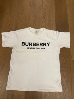 Vit Burberry t-shirt med tryck - Vit t-shirt från Burberry med klassiskt svart tryck 'BURBERRY LONDON ENGLAND' på bröstet. T-shirten har rund hals och korta ärmar, tillverkad i mjuk bomull för en skön och stilren look.