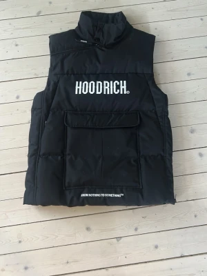 Svart dunväst från Hoodrich - Snygg svart dunväst från Hoodrich med stor ficka framtill och vit logga på bröstet. Västen har hög krage och trycket 'FROM NOTHING TO SOMETHING' längst ner. Perfekt för lager på lager och streetwear-stil.