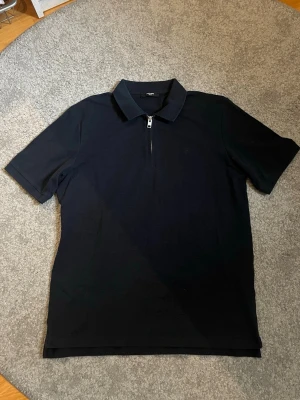 Marinblå Pikétröja  - Marinblå pikétröja från Jack & Jones med klassisk krage och half zip-dragkedja framtill. Toppen har korta ärmar och är tillverkad i ett mjukt material som känns skönt mot huden. Har endast använts 1-2 gånger, perfekt inför sommarsäsongen.