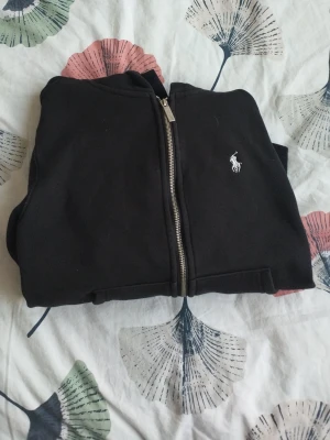 Svart hoodie från Polo Ralph Lauren - Klassisk svart hoodie från Polo Ralph Lauren med dragkedja framtill och den ikoniska vita logon broderad på bröstet. Hoodien har huva och är tillverkad i mjukt material som känns skönt mot huden. Perfekt för en avslappnad och stilren look.