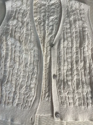 Vit virkad väst från NA-KD - Säljer en vit virkad väst från NA-KD i storlek M. Västen har ett snyggt hålmönster, ribbade kanter och dekorativa knappar framtill. Perfekt att styla över en t-shirt eller skjorta för en trendig lager-på-lager-look.