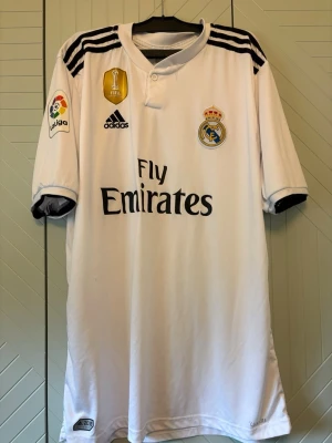 Real Madrid matchtröja Sergio Ramos - Vit Real Madrid matchtröja från Adidas med svarta detaljer, klubbmärke och sponsortryck på bröstet. Nummer 4 och Sergio Ramos på ryggen. La Liga-märke på ärmen och FIFA-märke på bröstet. Tillverkad i lätt och ventilerande material. Se sista bilden då den är lite trasig 