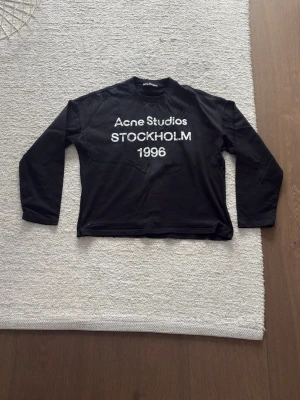Svart långärmad tröja Acne Studios - Svart långärmad tröja från Acne Studios med vit text 'Acne Studios STOCKHOLM 1996' på bröstet. Tröjan har rund hals och rak passform. Perfekt för en stilren och trendig look.