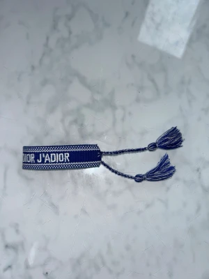 Blå armband  Dior - Snyggt blått tygarmband från Dior med texten 'J'ADIOR' och 'CHRISTIAN' i vitt. Armbandet har justerbara knytband med tofsar i ändarna och är perfekt för att ge din outfit en exklusiv touch.
