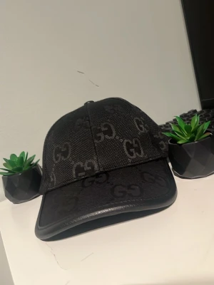 Svart keps från Gucci med GG-mönster - Snygg svart keps från Gucci med det klassiska GG-mönstret över hela kepsen. Kepsen har böjd skärm och är tillverkad i textil med detaljer i läder. Perfekt accessoar för en trendig och exklusiv look.