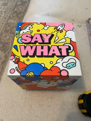 Sällskapsspel Say What?! - Säljer sällskapsspelet Say What?! från Ninja Print. Spelet har en färgglad box med tecknade figurer och text i rosa och svart. Det är ett charadspel med 1200 utmaningar där man gissar och förklarar ord i tre olika rundor. Perfekt för spelkvällar med vänner.