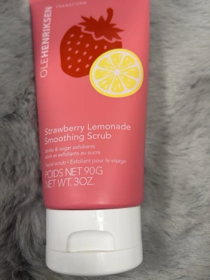 OLEHENRIKSEN Strawberry Lemonade Scrub - Ansiktsskrubb med doft av jordgubb och citron från OLEHENRIKSEN. Innehåller AHA och sockerexfolianter för att ge en mjukare och fräschare hud. Kommer i en rosa tub med vita detaljer och illustrationer av jordgubb och citron.