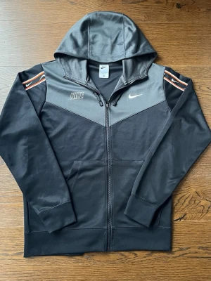 Svart och grå Nike hoodie med dragkedja - Nike hoodie i svart och grått med dragkedja framtill och huva. Snygga detaljer med vita och orange Nike-loggor på axlarna. Tillverkad i mjukt material, perfekt för en sportig och avslappnad stil.