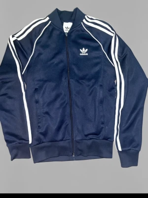 Mörkblå Adidas track jacket - Klassisk mörkblå Adidas track jacket med vita ränder längs ärmarna och vit Adidas-logga på bröstet. Jackan har dragkedja framtill, ribbade muddar och ståkrage. Perfekt för sport eller streetwear. Materialet är troligtvis polyester.