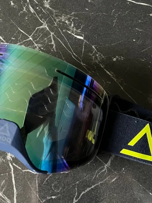 AMOQ skidglasögon med spegelglas  - Säljer ett par coola skidglasögon från AMOQ med spegelglas i blå och gröna nyanser. De har en bred, elastisk rem med gult AMOQ-tryck och vågigt silikonmönster på insidan för extra grepp. Perfekta för dig som vill sticka ut i backen! nypris 900kr
