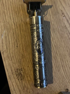 Metalltrimmer med Buddha-motiv - Snygg elektrisk hårtrimmer i silverfärgad metall med graverade detaljer och ett Buddha-huvud på handtaget. Trimmern har ett avlångt, cylinderformat grepp och ser riktigt stilren ut. Perfekt för dig som vill ha en unik och cool trimmer till håret eller skägget. Ingår laddare + extra trimmer kammar ”som sitter längst fram”