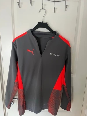 AC Milan träningströja från Puma (KÖPT FRÅN SAN SIRO) - Mörkgrå långärmad träningströja från Puma med röda detaljer och AC Milan-tryck på bröstet. Tröjan har halv dragkedja framtill, röda paneler på axlar och sidor samt mönstrade partier vid ärmslut. Perfekt för fotbollsträning eller sport. Den är köpt från SAN Siro!! Har någon defekt vid magen typ men inget hål som syns så mycket! 