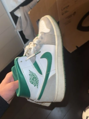 Air Jordan 1 Mid vit/grön - Snygga Air Jordan 1 Mid sneakers i vitt och grönt med klassisk vinge-logga på sidan. Ovandel i läder och mocka, gröna detaljer på swoosh, häl och innerfoder. Perforerad tåbox för extra ventilation och cool look. Perfekta för dig som gillar streetwear och basketstil.