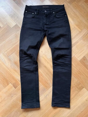 Svarta slim jeans från Nudie Jeans - Snygga svarta slim fit jeans från Nudie Jeans med klassisk femficksdesign och raka ben. Jeansen har diskreta nitar och knappgylf, samt Nudie-logga på bakfickan. Perfekta för en stilren och modern look. Modell Grim Tim. W:31 L:32