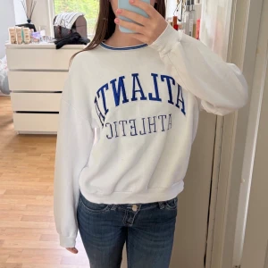 Vit sweatshirt med blå text - Vit oversized sweatshirt med blå text 'ATLANTIC ATHLETIC' på bröstet och en stor blå siffra 8 på ena ärmen. Tröjan har rund halsringning med blå detalj och avslappnad passform. Perfekt för en sportig och chill look. Lite skavanker som ni kan se på det sista 2 bilderna därav billigare pris! Vet ej märket