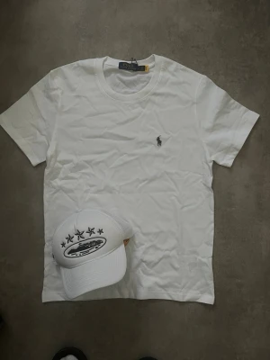 Vit t-shirt från Polo Ralph Lauren - Vit t-shirt från Polo Ralph Lauren med klassisk rund hals och ikoniska broderade loggan på bröstet. Enkel och stilren design som passar till det mesta. Tillverkad i mjuk bomull för en skön känsla.
