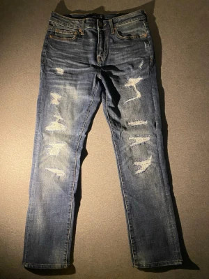 Blå slitna jeans med raka ben - Säljer ett par blå jeans med raka ben och tydliga slitningar framtill. Jeansen har klassisk femficksdesign och är tillverkade i denim. Perfekta för en avslappnad och trendig look.