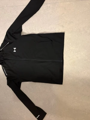 Under Armour vanish - Snygg svart träningsjacka från Under Armour med dragkedja framtill och vit logga på bröstet. På ryggen står det 'UNDER ARMOUR' i vitt. Jackan har långa ärmar och är tillverkad i ett lätt, funktionellt material som passar perfekt till träning och löpning.