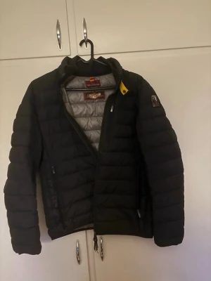 Svart pufferjacka från Parajumpers - Snygg svart pufferjacka från Parajumpers med quiltad design och grått innerfoder. Jackan har dragkedja, bröstficka med dragkedja och klassisk gul detalj vid kragen. Märkespatch på ärmen och lätt vadderad för extra värme.