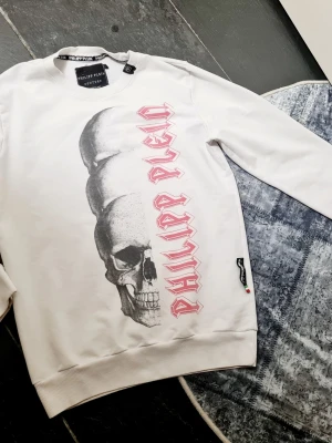 Vit sweatshirt från Philipp Plein - Säljer en vit sweatshirt från Philipp Plein med en stor dödskalle-grafik och rosa text längs framsidan. Tröjan har rund halsringning, långa ärmar och ribbade muddar. Materialet är mjukt och bekvämt, perfekt för en edgy streetstyle-look.