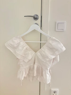 Vit volangblus med snörning - Supersöt vit blus med volanger och v-ringning. Toppen har snörning i ryggen och är ärmlös med breda volangdetaljer över axlarna. Perfekt för en somrig och romantisk look.