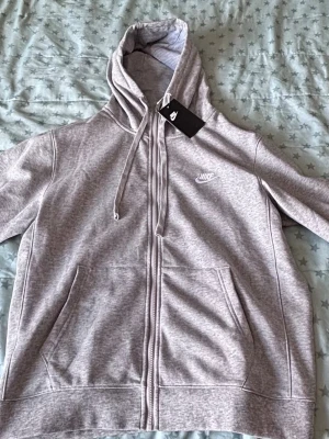 Grå zip-hoodie från Nike - Snygg gråmelerad hoodie från Nike med dragkedja, huva med dragsko och två stora fickor framtill. Klassisk Nike-logga broderad på bröstet. Mjuk bomullsblandning som är skön att ha på sig. Perfekt för en avslappnad streetstyle.