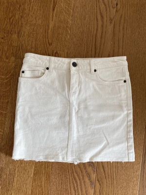 Vit Lexington jeanskjol med rå kant - Snygg vit jeanskjol från Lexington med klassisk femficksdesign, bälteshällor och rå kant nertill. Kjolen har knapp och dragkedja framtill samt en liten läderpatch med logga bak i midjan. Aldrig använd.