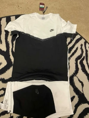 Nike svartvitt set med t-shirt & shorts - Snyggt svartvitt set från Nike med t-shirt och matchande shorts. T-shirten har en vit överdel och svart underdel med Nike-logga på bröstet. Shortsen är vita med svart panel och Nike-logga. Perfekt för en sportig och avslappnad look.