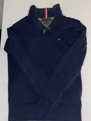 Marinblå halvzip-tröja Tommy Hilfiger - Klassisk marinblå stickad tröja från Tommy Hilfiger med halv dragkedja och ståkrage. Tröjan har långa ärmar och diskret logga på bröstet. Perfekt för en stilren och avslappnad look.