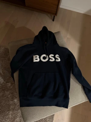 Mörkblå hoodie från BOSS - Snygg mörkblå hoodie från BOSS med stor vit logga på bröstet. Klassisk modell med huva och dragsko samt en stor magficka framtill. Perfekt för en avslappnad och trendig stil. Mjukt material som känns skönt mot huden.