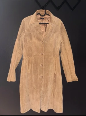 Beige mockakappa med knappar - Stilren beige kappa i mocka med klassisk krage och knäppning framtill. Lång modell med markerad midja och diskreta fickor. Perfekt för dig som gillar en tidlös och trendig look.
