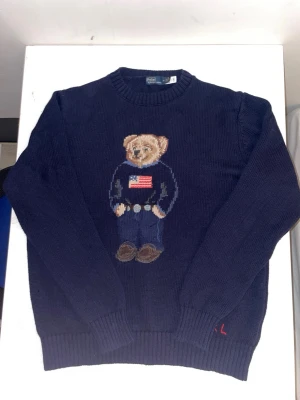 Mörkblå stickad tröja Polo Bear - Tröjan är använd i en vecka. Mörkblå stickad tröja från Polo Ralph Lauren med den ikoniska Polo Bear på bröstet, klädd i jeans och tröja med amerikanska flaggan. Rund halsringning och ribbade muddar vid ärmslut och nederkant. RL-broderi nertill.