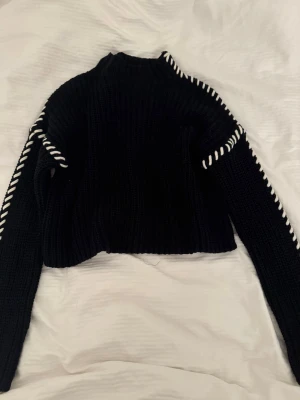 Svart stickad tröja från Vero Moda - Snygg svart stickad tröja från Vero Moda med vita detaljer längs ärmar och axlar. Tröjan har en croppad passform, hög hals och är ribbstickad för extra struktur. Perfekt för att lyfta din outfit med en cool kontrast. 