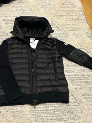 Svart pufferjacka från Moncler - Snygg svart pufferjacka från Moncler med quiltad front och huva. Ärmar och rygg i mjukt stickat material, dragkedja framtill och två fickor med dragkedja. Moncler-logga på ena ärmen. Perfekt för kalla vinterdagar.