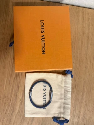 Blått armband från Louis Vuitton - Snyggt armband från Louis Vuitton i blått textilmaterial med silvriga metalldetaljer. Kommer med original ask och dustbag. Perfekt accessoar för att lyfta din outfit med en exklusiv touch.