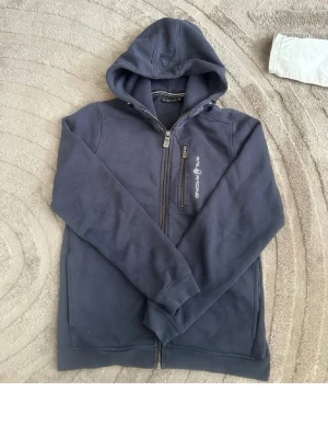 Mörkblå hoodie från Sail Racing - Mörkblå hoodie med dragkedja från Sail Racing. Har huva med dragsko, bröstficka med dragkedja och Sail Racing-logga på bröstet. Mjuk bomullsmix och två sidofickor. Perfekt för en avslappnad och sportig stil.