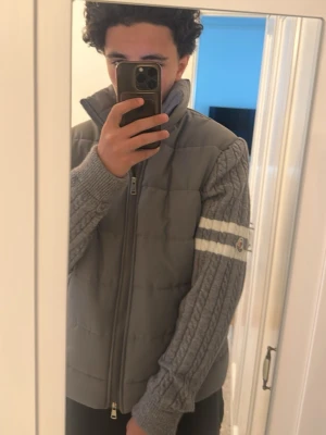 Grå cardiganjacka från Moncler - Snygg grå cardiganjacka från Moncler med stickade ärmar och quiltad front. Jackan har dragkedja, högt krage och vita ränder på ena ärmen samt Moncler-logga. Perfekt för lager-på-lager och streetwear-vibe.OBS! Den är storlek XL men passar L