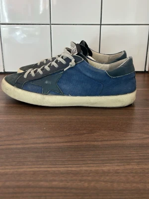 Golden goose sneakers - Snygga blå sneakers från Golden Goose med grå detaljer och vit sula. Skorna har en ikonisk stjärndesign på sidan, gråa skosnören och ovandel i canvas och mocka. Perfekta för dig som gillar en avslappnad och trendig stil.