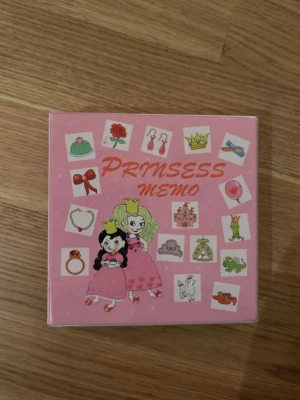 Prinsess Memo spel rosa - Ett memoryspel med prinsesstema i rosa ask. Innehåller brickor med motiv som kronor, smycken, klänningar och djur. Perfekt för att träna minnet och ha kul med vänner eller familj. Sött och färgglatt designat för prinsessfantaster.