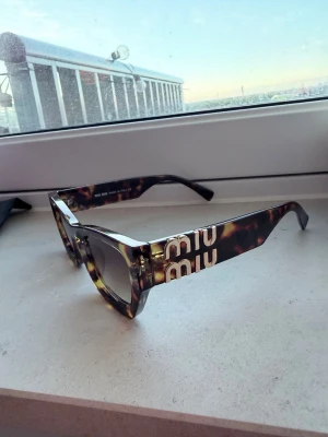Miu Miu solglasögon i leopardmönster - Snygga solglasögon från Miu Miu med trendigt leopardmönster och stora bågar. Modellen har en markerad cat-eye form och logotypen i vitt på sidorna. Perfekta för att lyfta din stil med en cool och lyxig touch.