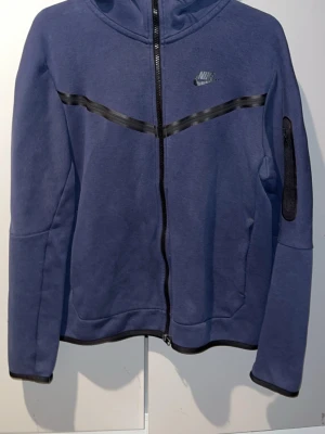 Blå kofta med dragkedja från Nike - Snygg blå hoodie från Nike med hel dragkedja och svart Nike-logga på bröstet. Hoodien har en svart detalj över bröstet och ärmen samt fickor på sidorna. Perfekt för en sportig och avslappnad stil. Storlek M 