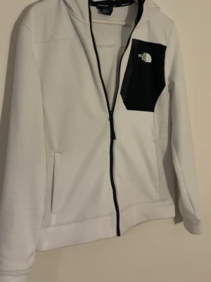 Vit hoodie från The North Face - Vit hoodie med svart bröstficka och dragkedja från The North Face. Hoodien har huva, långa ärmar och logga på både bröstet och axeln. Materialet är mjukt och stretchigt, perfekt för en sportig och clean stil.