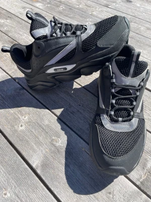 sneakers b22 med mesh och reflex - Snygga svarta sneakers med coola reflexdetaljer och meshpaneler för extra andningsförmåga. Skorna har en chunky sula och sportig design med grå och silvriga inslag. Perfekta för dig som vill sticka ut med en exklusiv streetstyle.