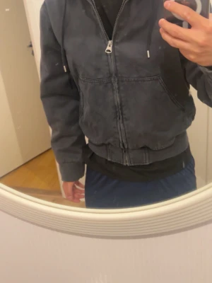 Mörkgrå hoodie med dragkedja H&M - Säljer en mörkgrå hoodie från H&M med dragkedja framtill och två stora fickor. Modellen har huva med dragsko och ribbade muddar vid ärmslut och nederkant. Perfekt för en avslappnad och stilren look.