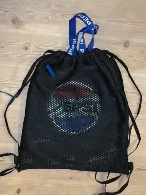 Svart mesh gympåse från Pepsi - Cool svart gympåse i meshmaterial med stort Pepsi-tryck på framsidan och blått band med Pepsi-logga. Dragskostängning och justerbara axelband. Perfekt för träning eller som streetstyle-accessoar.