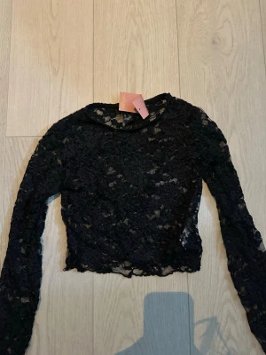 Svart spetsblus från Refuge - Snygg svart långärmad blus från Refuge i genombruten spets med blommigt mönster. Croppad modell som ger en trendig look. Perfekt att styla med höga jeans eller kjol för en cool outfit.