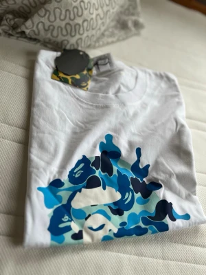 Vit BAPE t-shirt med blå camo-tryck - Snygg vit t-shirt från BAPE med ett stort blått camo-tryck framtill. Gjord i 100% bomull och har klassisk passform med rund hals. Perfekt för dig som gillar streetwear och vill sticka ut med en unik design.