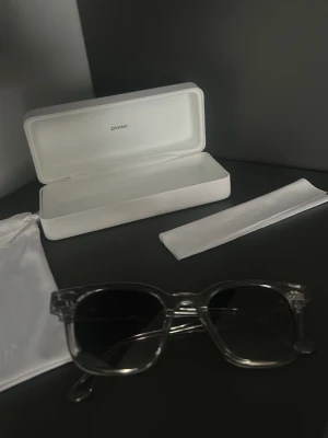 Chimi 02  - Säljer ett par stilrena solglasögon från Chimi med transparent båge och mörka glas. Medföljer vit hård förvaringsbox. tygpåse samt putsduk. Perfekt accessoar för dig som vill ha en clean och modern look. Finns en repa därav priset annars väldigt fräscha 