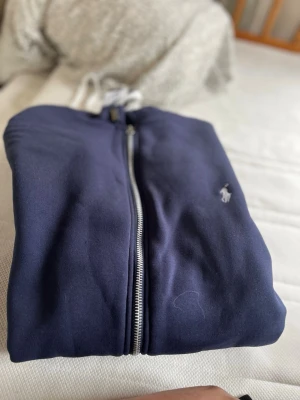 Marinblå hoodie från Polo Ralph Lauren - Snygg marinblå hoodie från Polo Ralph Lauren med dragkedja framtill och den klassiska broderade loggan på bröstet. Hoodien har en enkel och stilren design, perfekt för en avslappnad look. Tillverkad i mjuk bomull för extra komfort.